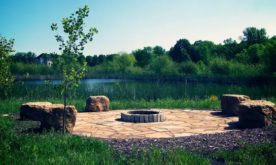 Circular stone fire pit on a flagstone patio with boulder seating overlooking a pond at dusk