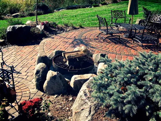 Circular brick paver patio with a boulder-ringed fire pit and wrought-iron seating beside a landscaped border