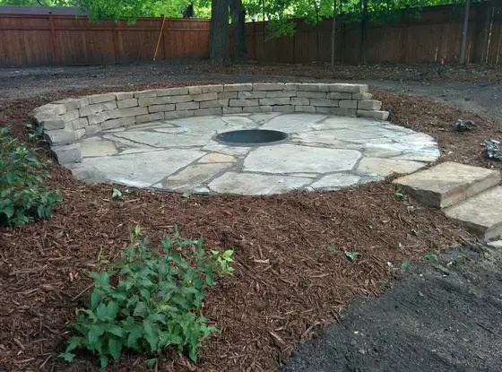 Flagstone fire pit patio with a curved stacked-stone seating wall and mulched garden beds in a wooded backyard