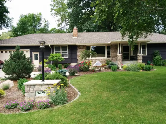 Edina rambler with a stone address pillar, colorful perennial beds, and boulder-edged foundation plantings
