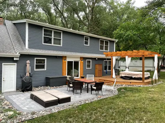 Backyard paver patio with a cedar pergola, hot tub, and outdoor dining set behind a gray two-story home