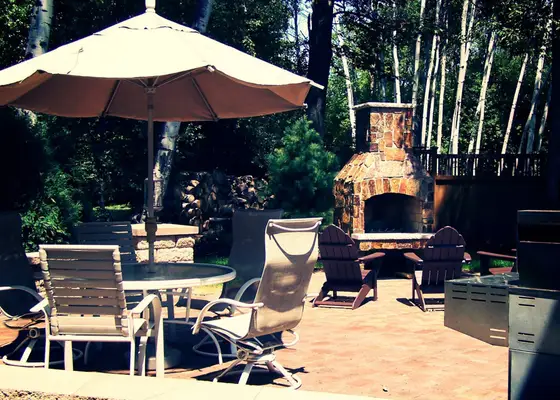 Paver patio outdoor living space with umbrella dining set, stacked-stone fireplace, and built-in grill among evergreens