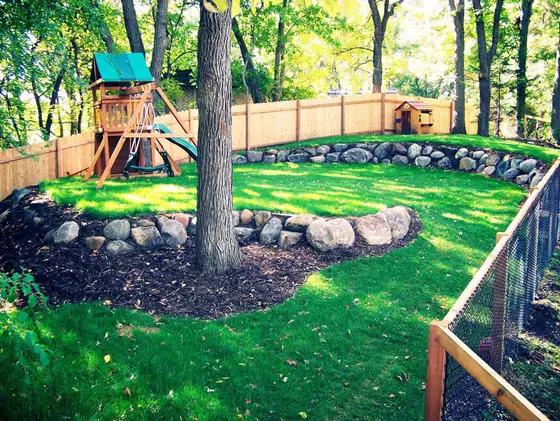 Backyard boulder retaining wall edging a manicured lawn with a cedar privacy fence and playset under a mature tree