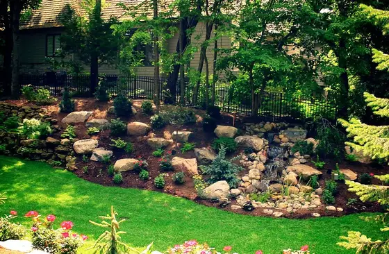 Boulder-lined backyard water feature surrounded by perennial plantings and a manicured lawn near an iron fence